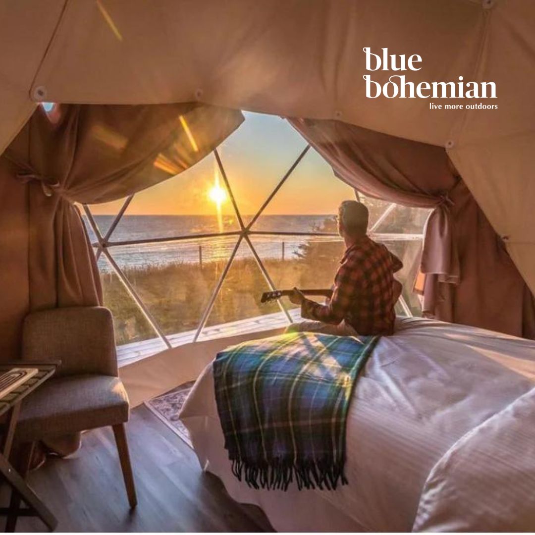 Person sitting on a bed in a glamping dome with a scenic view and 'blue bohemian' branding.