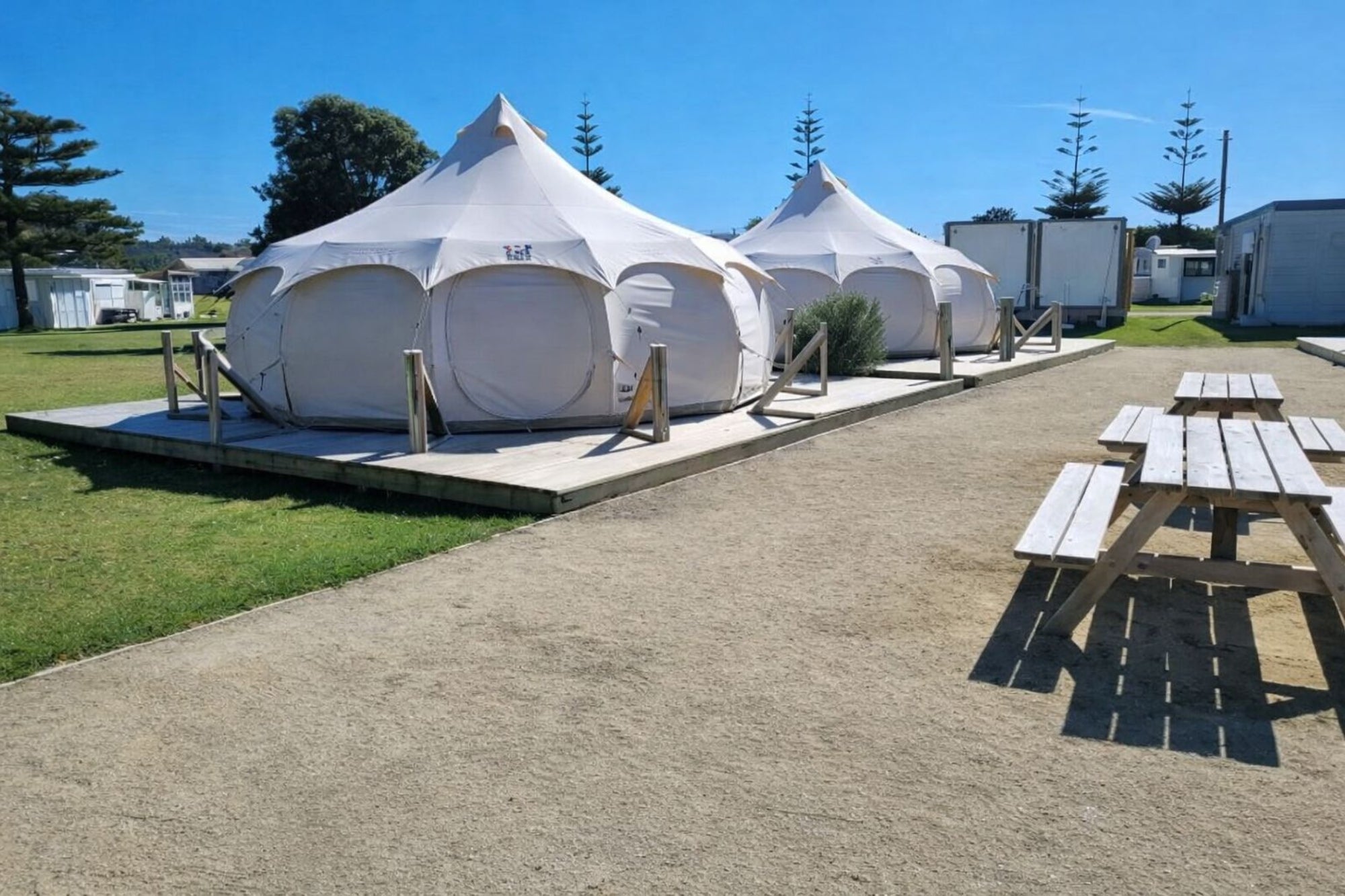 Two Better Bell yurt tents set up on wooden decks in a coastal camping area, featuring distinct arched windows and conical peaks under a clear blue sky.
